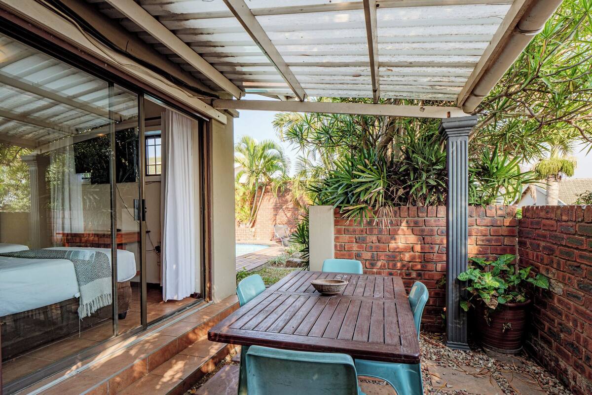 A covered patio with a wooden table and chairs, a glass door leading to a bedroom, and a brick wall with greenery.