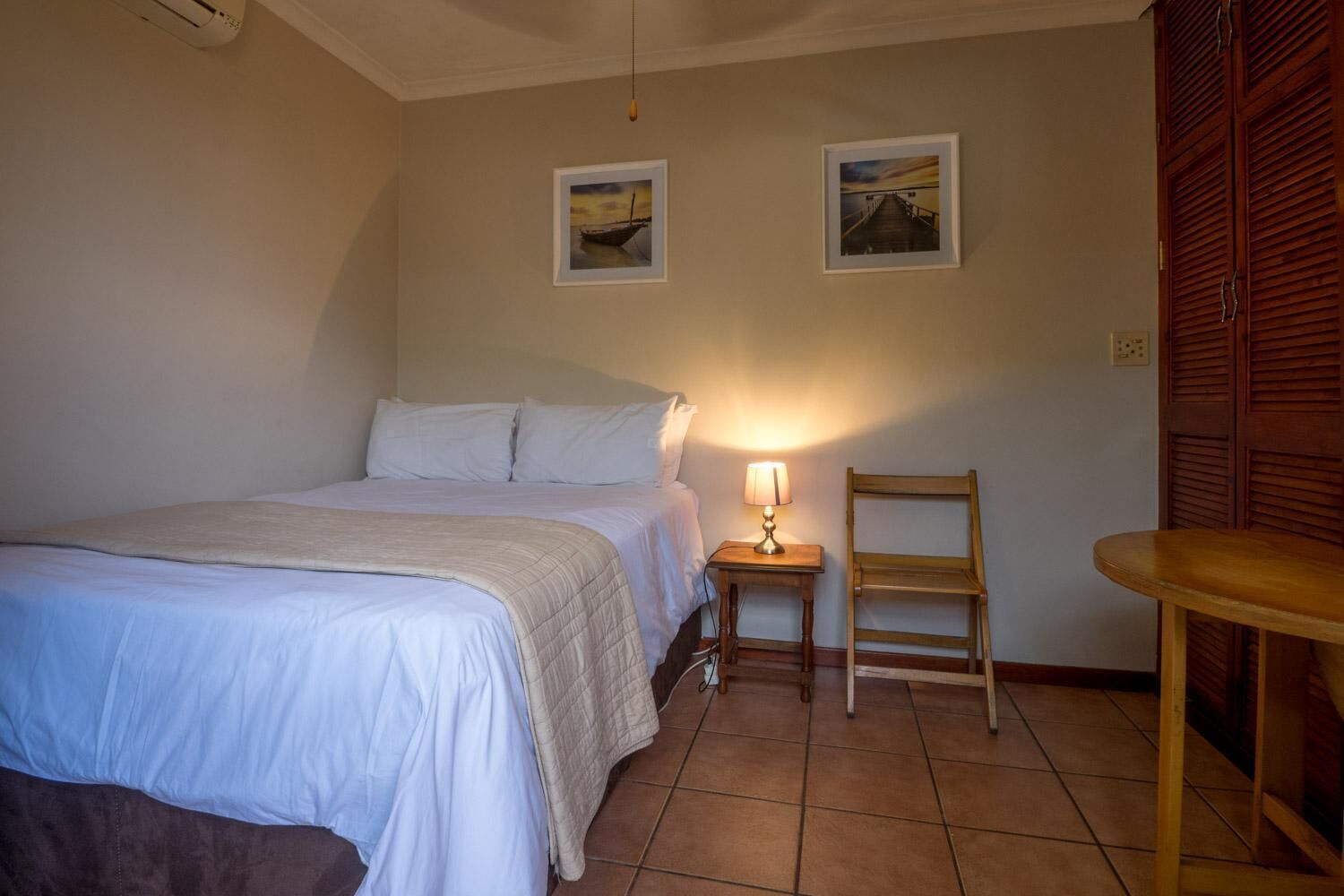 Un dormitorio con una cama, una mesita de noche con lámpara, una silla y dos cuadros en la pared.