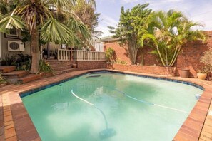 A swimming pool with a brick border, surrounded by palm trees and a brick wall. - Carrington Guest House (Durban)