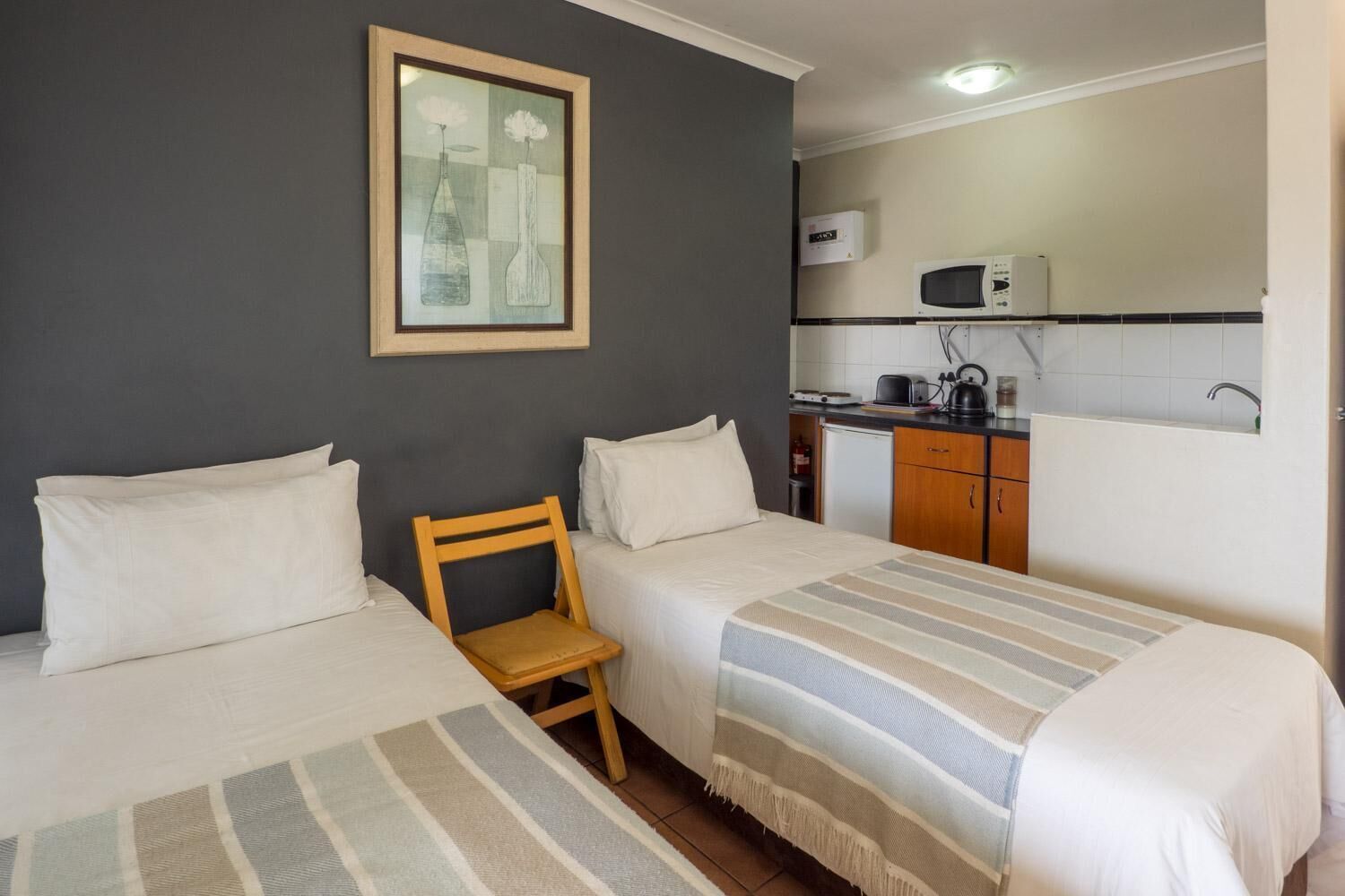 Habitación de hotel con dos camas, una pequeña silla de madera, una kitchenette con microondas, tostadora, hervidor y un lavabo.