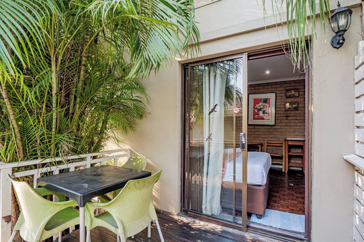 A patio with a table and chairs, a glass door leading to a room with a bed, and a large plant.