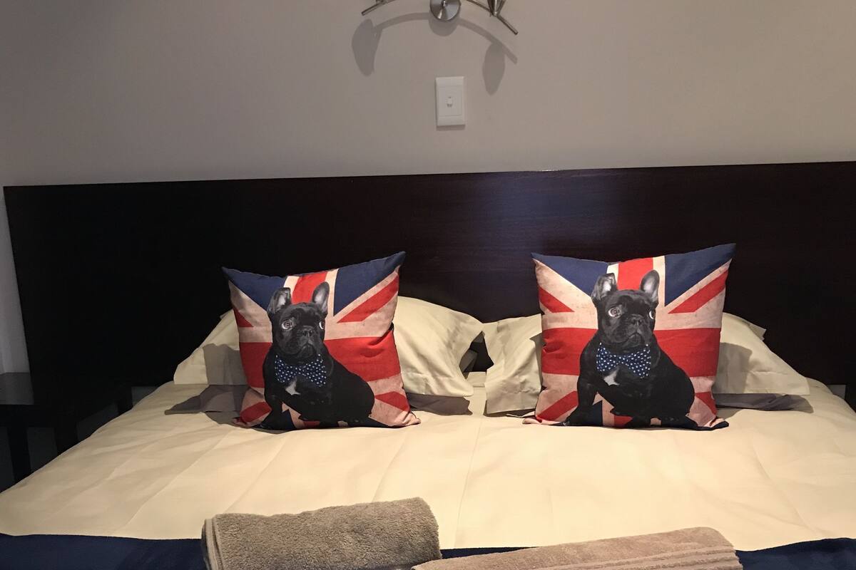 A bed with two pillows featuring a dog and a Union Jack design, a headboard, and a wall-mounted light fixture.