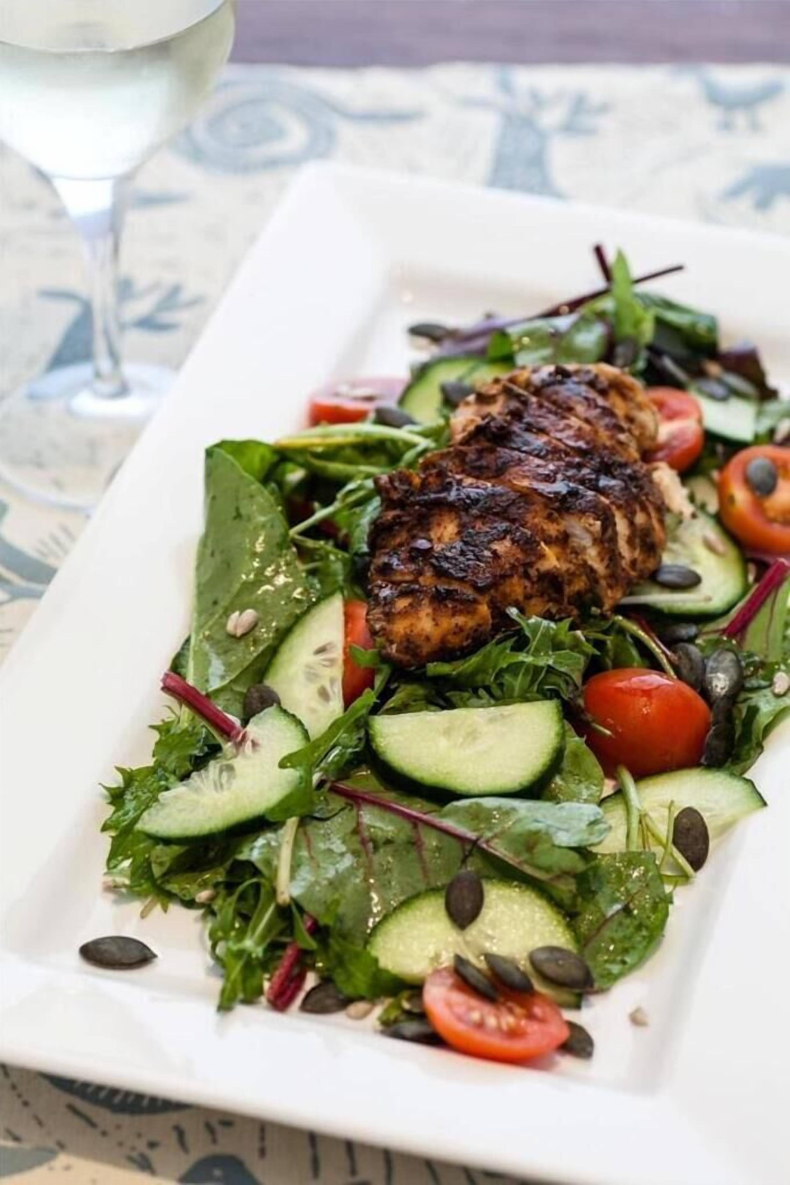 Un plato rectangular blanco con pechuga de pollo a la parrilla, ensalada mixta, rodajas de pepino, tomates cherry y pimienta negra.