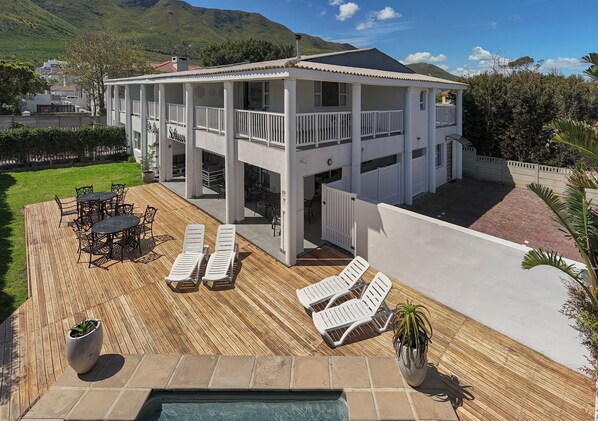 Una casa de dos pisos con terraza de madera, mobiliario exterior y piscina.
