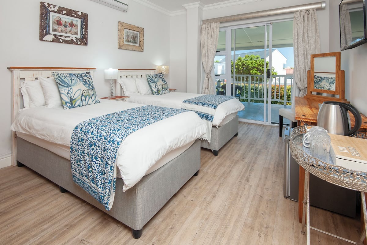 A hotel room with two beds, a balcony, a desk with a kettle, and a view of the outside.
