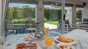 Mesa posta com itens de café da manhã, como ovos, bacon e torradas, com vista para um jardim e área de estar externa.