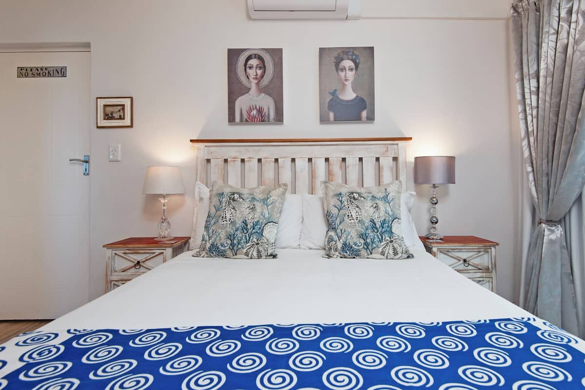A bedroom with a bed, two wall-mounted paintings, a bedside table with a lamp, and a "NO SMOKING" sign on the door.