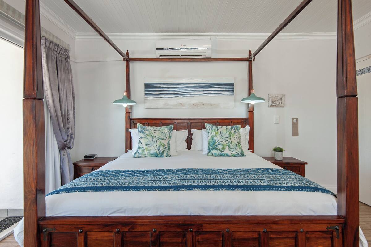 A four-poster bed with a wooden frame, a blue and white patterned bedspread, and two matching pillows. There is a wall-mounted air conditioning unit above the bed, a framed artwork depicting waves, and a small potted plant on the nightstand.