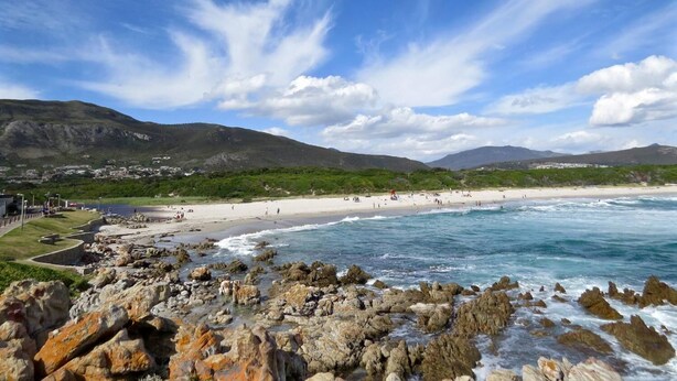 Uma praia costeira com pedras em primeiro plano, céu claro e montanhas ao longe.