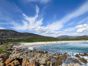 A coastal beach with rocky foreground, clear skies, and distant mountains. - Oude Schuur Boutique (Hermanus)