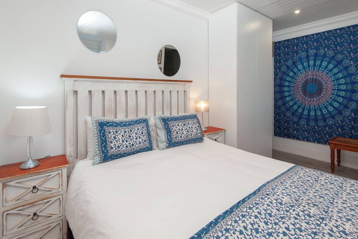 A bedroom with a white bed, two patterned pillows, a round mirror, a bedside table with a lamp, and a tapestry on the wall.