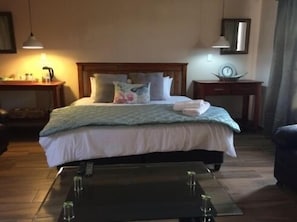 A neatly made bed with pillows, a bedside table with a lamp, and a small table with glasses. - Cura Lodge (Bloemfontein)
