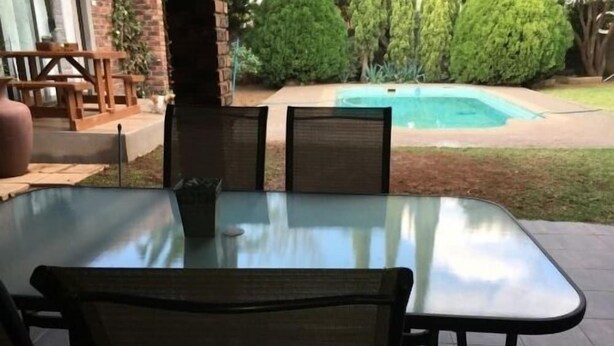 Um pátio externo com mesa e cadeiras de vidro, parede de tijolos e vista para uma piscina.
