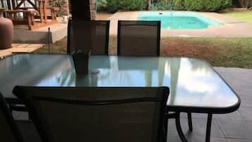 An outdoor patio with a glass table and chairs, a brick wall, and a pool in the background.