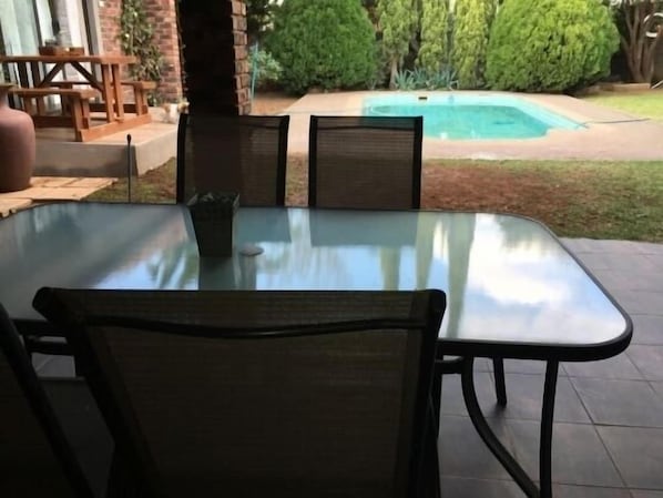 An outdoor patio with a glass table and chairs, a brick wall, and a view of a swimming pool. - Cura Lodge (Bloemfontein)