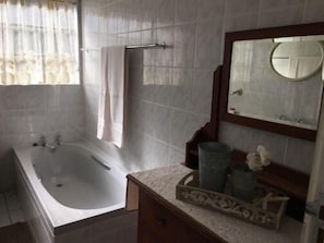 A bathroom with a bathtub, a vanity with a mirror, and a window with curtains. - Cura Lodge (Bloemfontein)