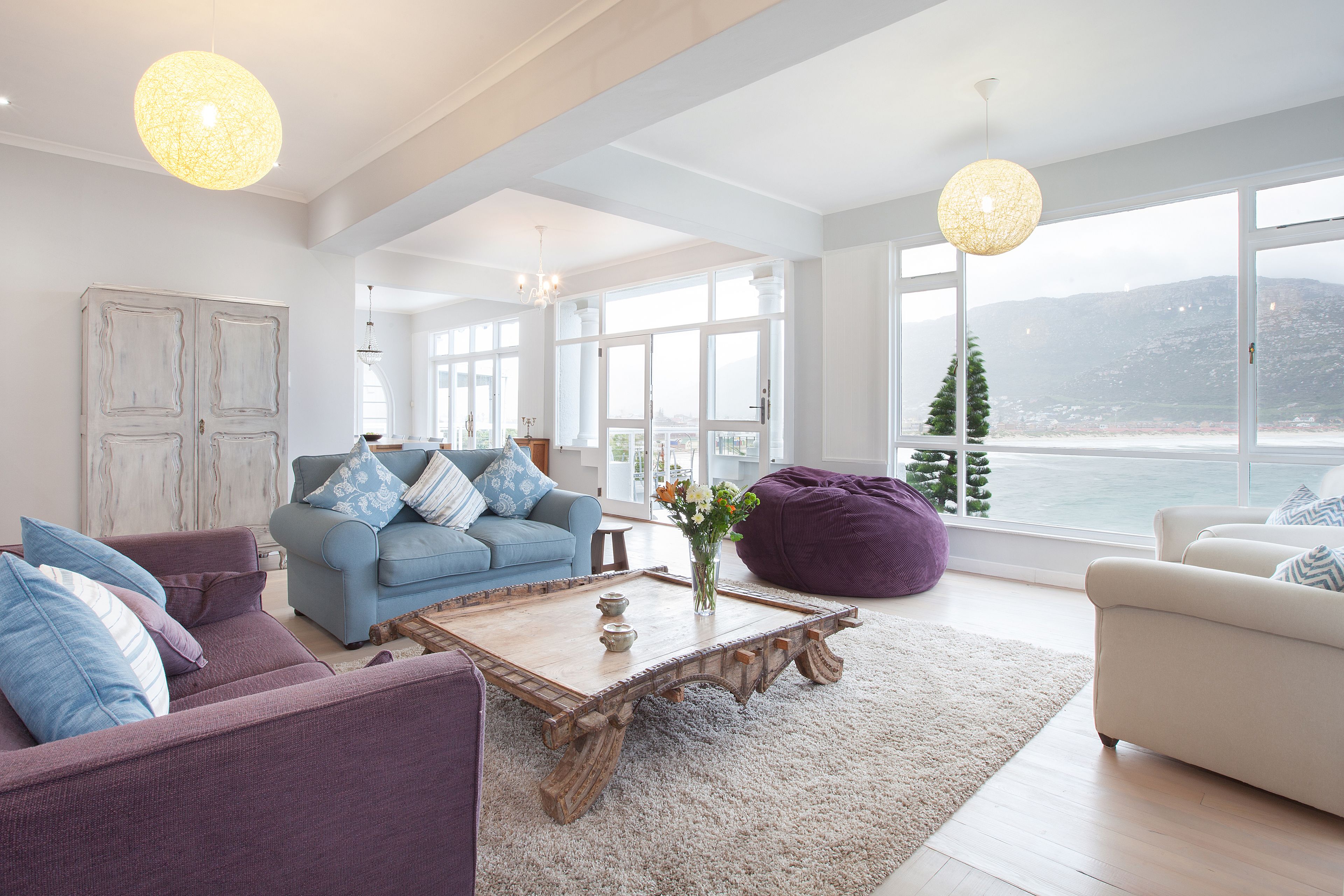 Uma sala de estar ampla com uma grande janela que oferece vista para o mar e montanhas, uma mesa de centro de madeira e móveis modernos.