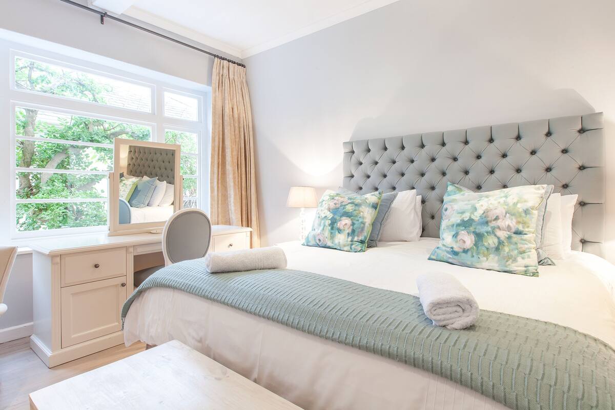 A bedroom with a large bed, a bedside table with a lamp, a window with curtains, and a mirror.