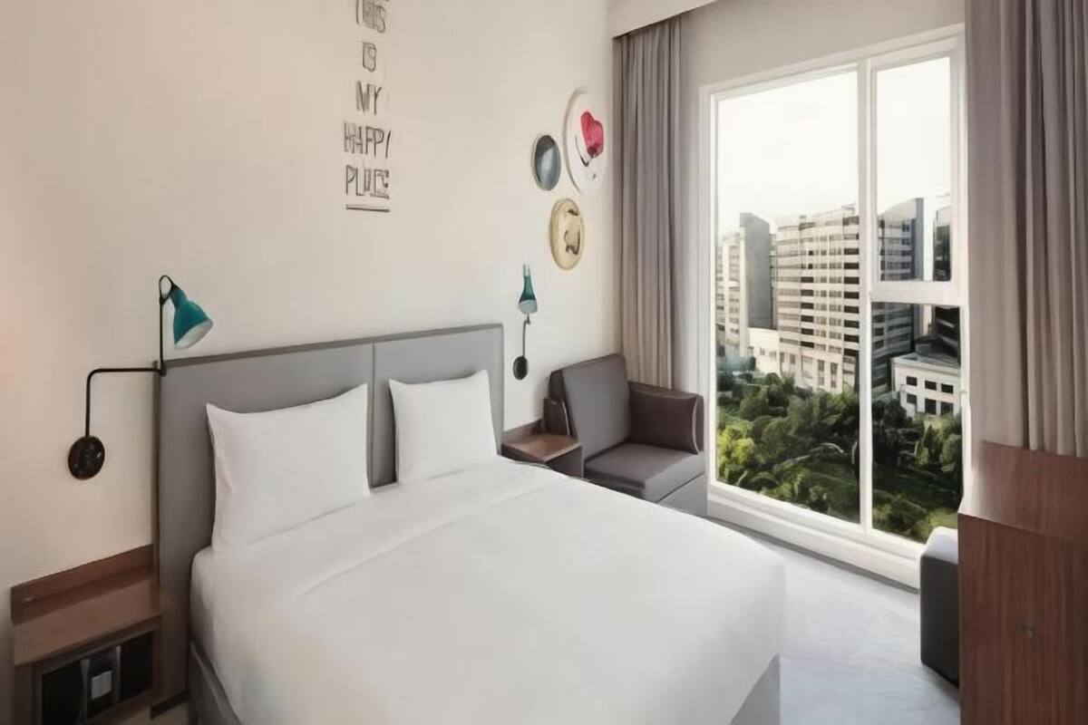 A hotel room with a large bed, a desk, a chair, a window with a city view, and a wall with a motivational quote.