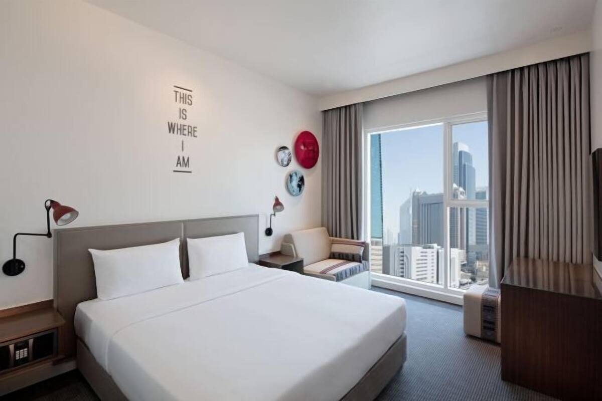 A hotel room with a large bed, a desk, a chair, a TV, and a window with a city view.