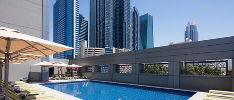 A rooftop pool with sun loungers and umbrellas, surrounded by high-rise buildings.