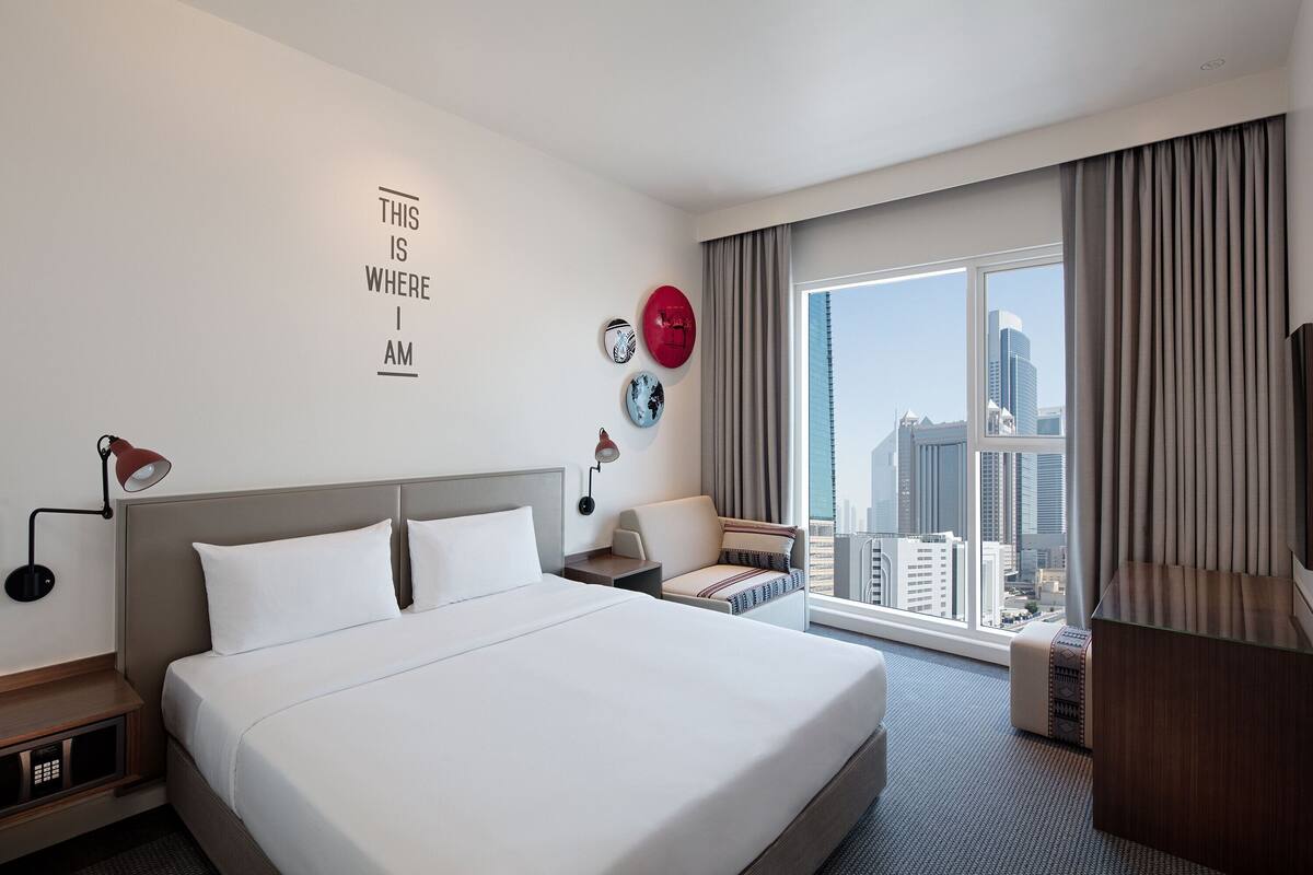 A hotel room with a large bed, a desk, a chair, a TV, and a view of the city.