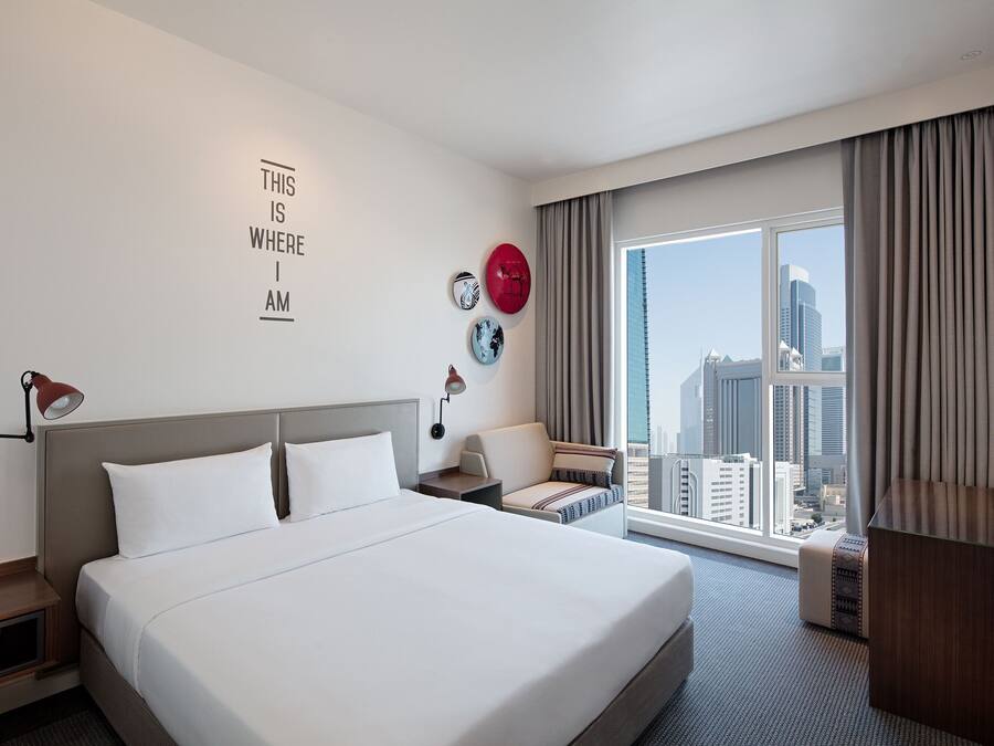 Quarto de hotel com uma cama grande, uma escrivaninha, uma cadeira, uma televisão e vista para a cidade.