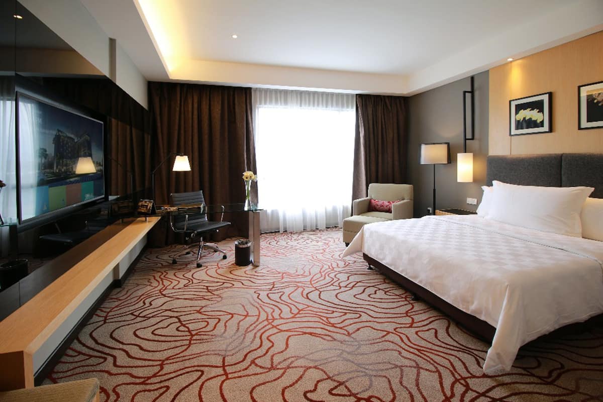 Une chambre d’hôtel avec un grand lit, un bureau avec un ordinateur, une télévision et une grande fenêtre avec des rideaux.