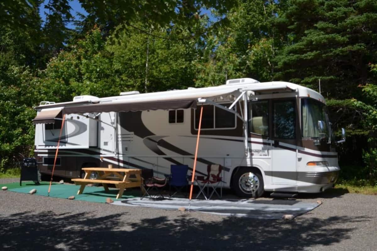 A recreational vehicle parked in a wooded area with a picnic table and chairs nearby.