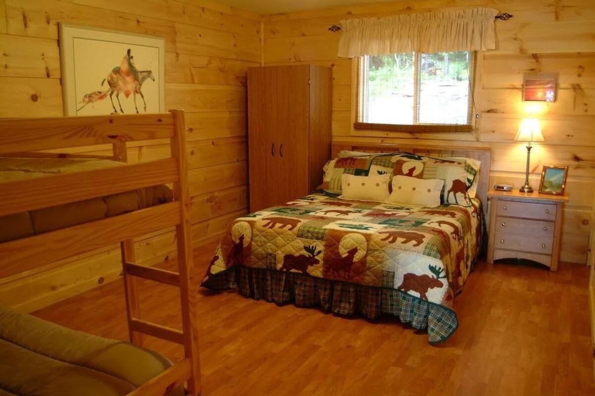 A cozy cabin room with a bunk bed, a quilted bed with animal prints, a wooden wardrobe, a window with curtains, and a bedside table with a lamp.