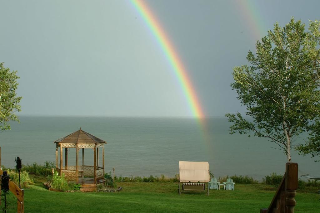 Un belvédère, une chaise couverte et un arbre au bord d’un plan d’eau, avec un arc-en-ciel en arrière-plan.