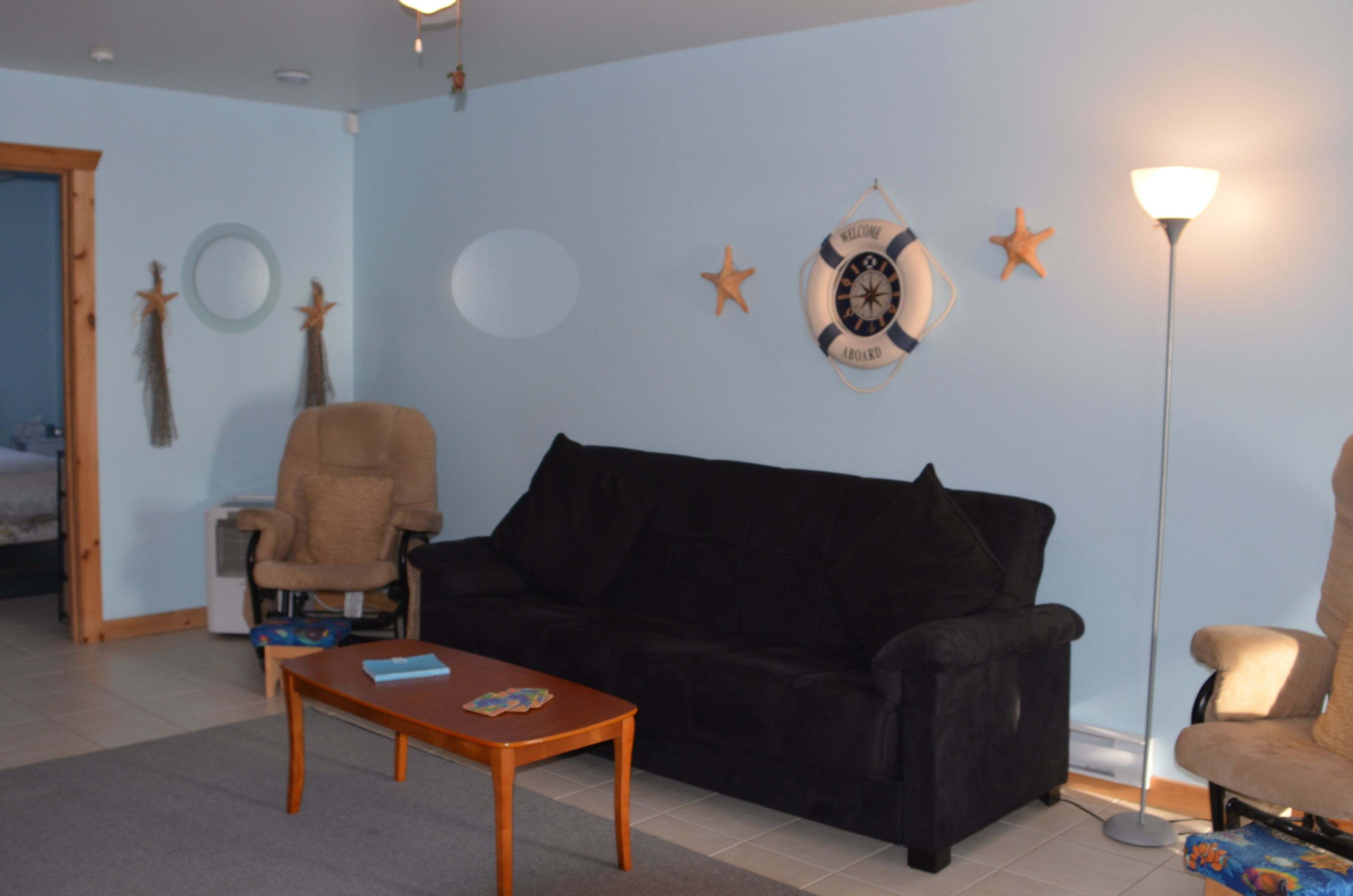 Ein Wohnzimmer mit einer schwarzen Couch, einem Holztisch und einem beige farbenen Sessel. An der Wand befinden sich Dekorationen mit Seesternen und ein runder Spiegel.