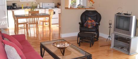 A living room with a glass coffee table, a red sofa, a black leather chair, a television, and a kitchen in the background.