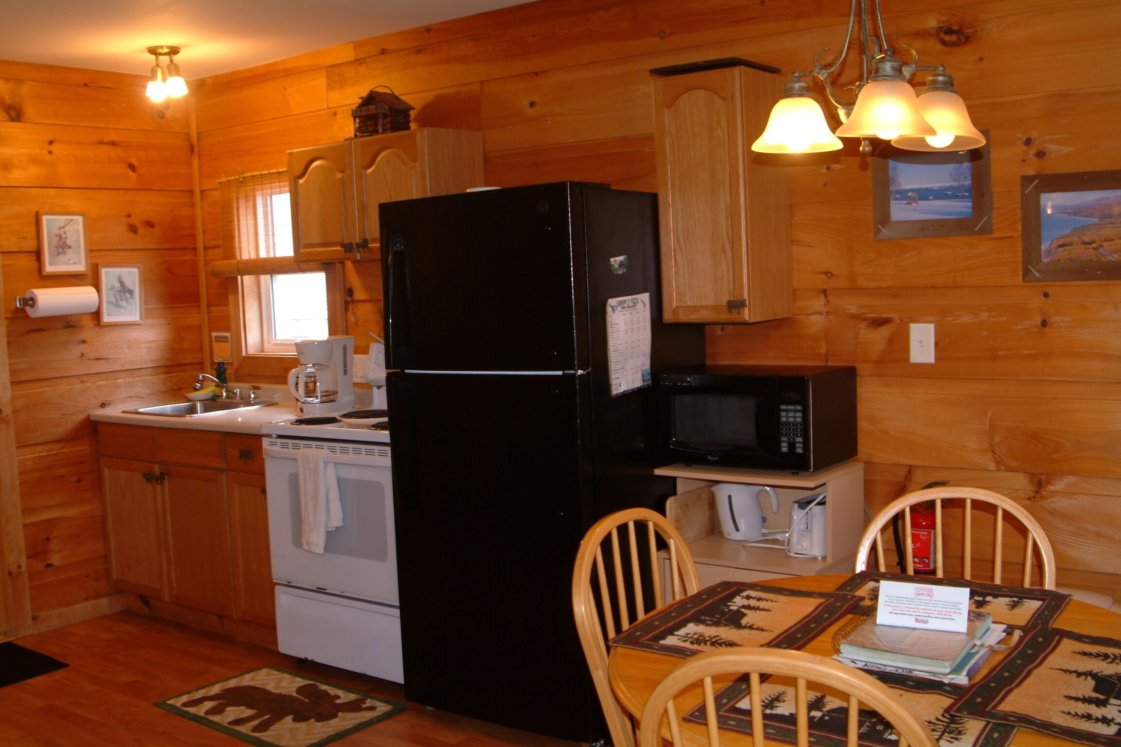 Cocina de cabaña de madera con un refrigerador negro, estufa blanca y gabinetes de madera. Hay una mesa de comedor con sillas y una pequeña isla con microondas y cafetera.