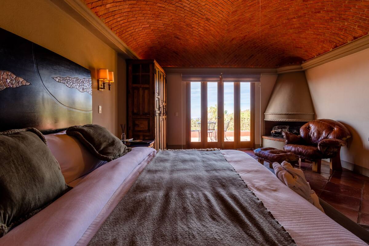 A bedroom with a large bed, a leather armchair, a television, and a fireplace.