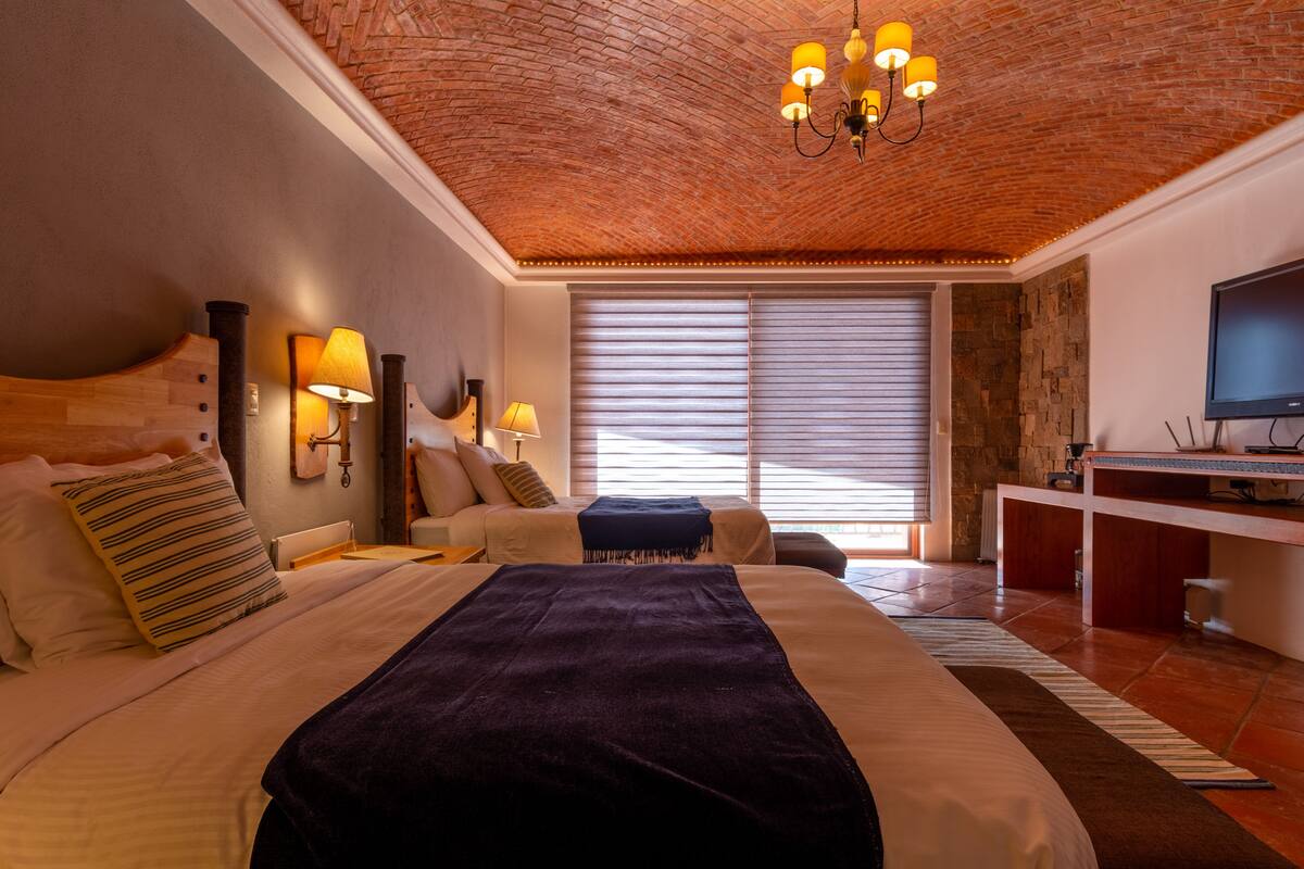 A modern hotel room with a brick ceiling, a large bed with a dark throw, a desk with a flat-screen TV, and a large window with blinds.