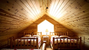 Quarto de sótão aconchegante com paredes de madeira e teto inclinado, com duas camas e uma mesinha.