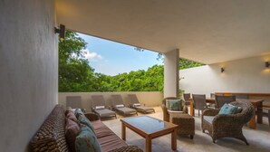 A covered outdoor seating area with wicker furniture, a glass-top table, and a view of lush greenery. - Magnificent 3BR condo close to the Caribbean sea by Happy Ad (Playa del Carmen)