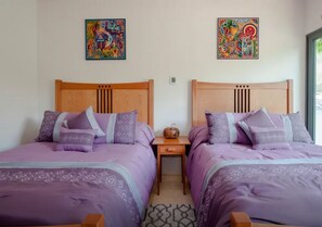 Two twin beds with matching headboards and purple bedding, a small bedside table with a decorative item, and colorful artwork on the walls. - Magnificent 3BR condo close to the Caribbean sea by Happy Ad (Playa del Carmen)