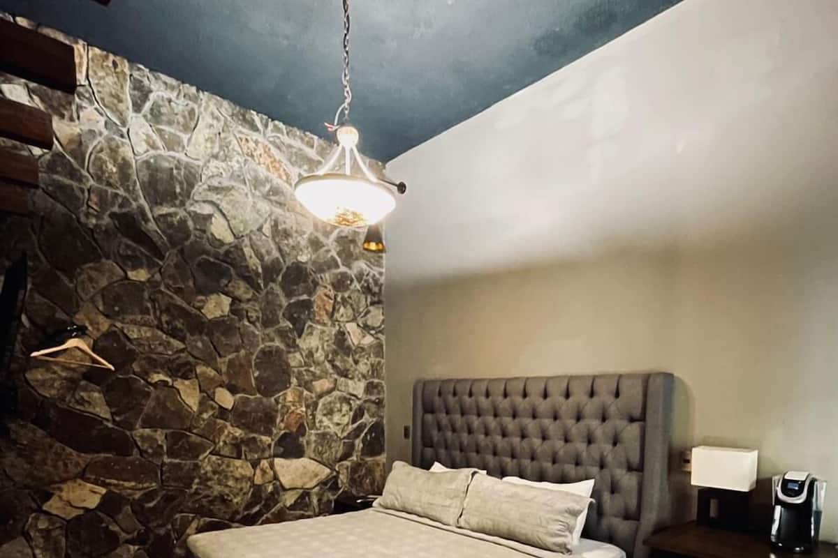 Une chambre avec un mur en pierre, un lit doté d’une tête de lit capitonnée, un plancher en bois, un luminaire suspendu et une petite table avec une lampe et une machine à café.