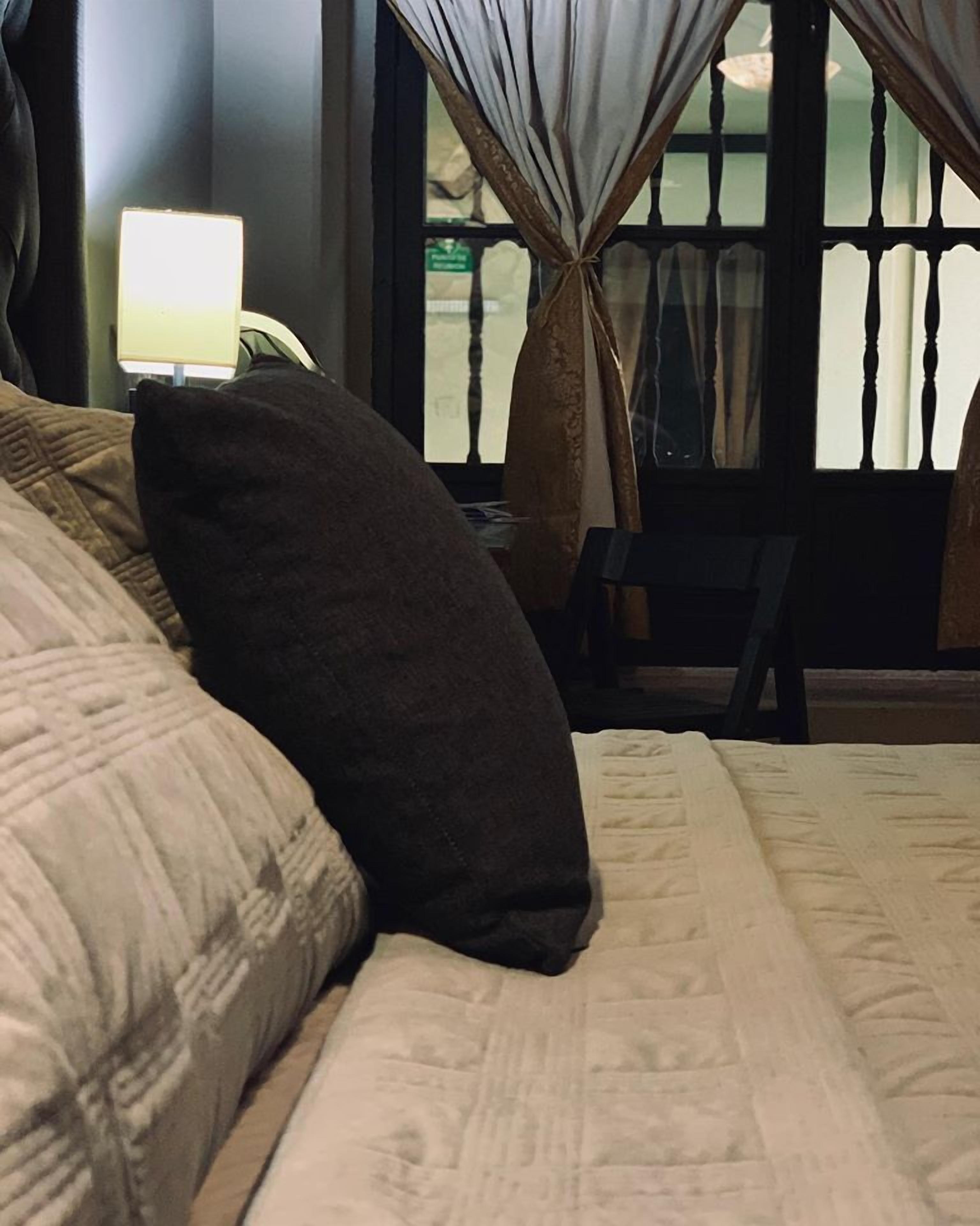 A neatly made bed with a dark pillow and a patterned blanket, a bedside lamp, and a window with curtains.