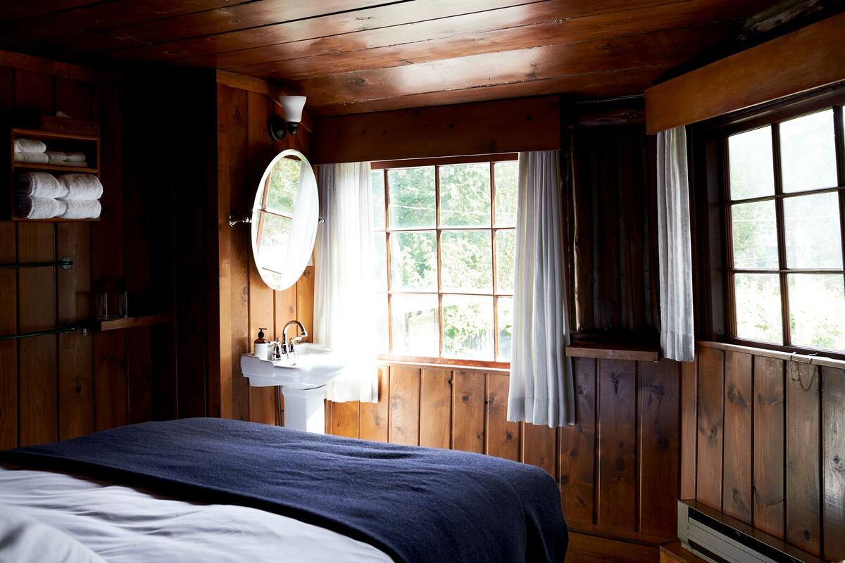 A wooden cabin bedroom with a bed, a window with curtains, a sink with a mirror, and a shelf with towels.