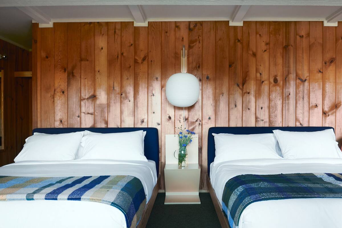 A room with two beds, wooden panel walls, a white hanging light fixture, and a small table with a vase of flowers.