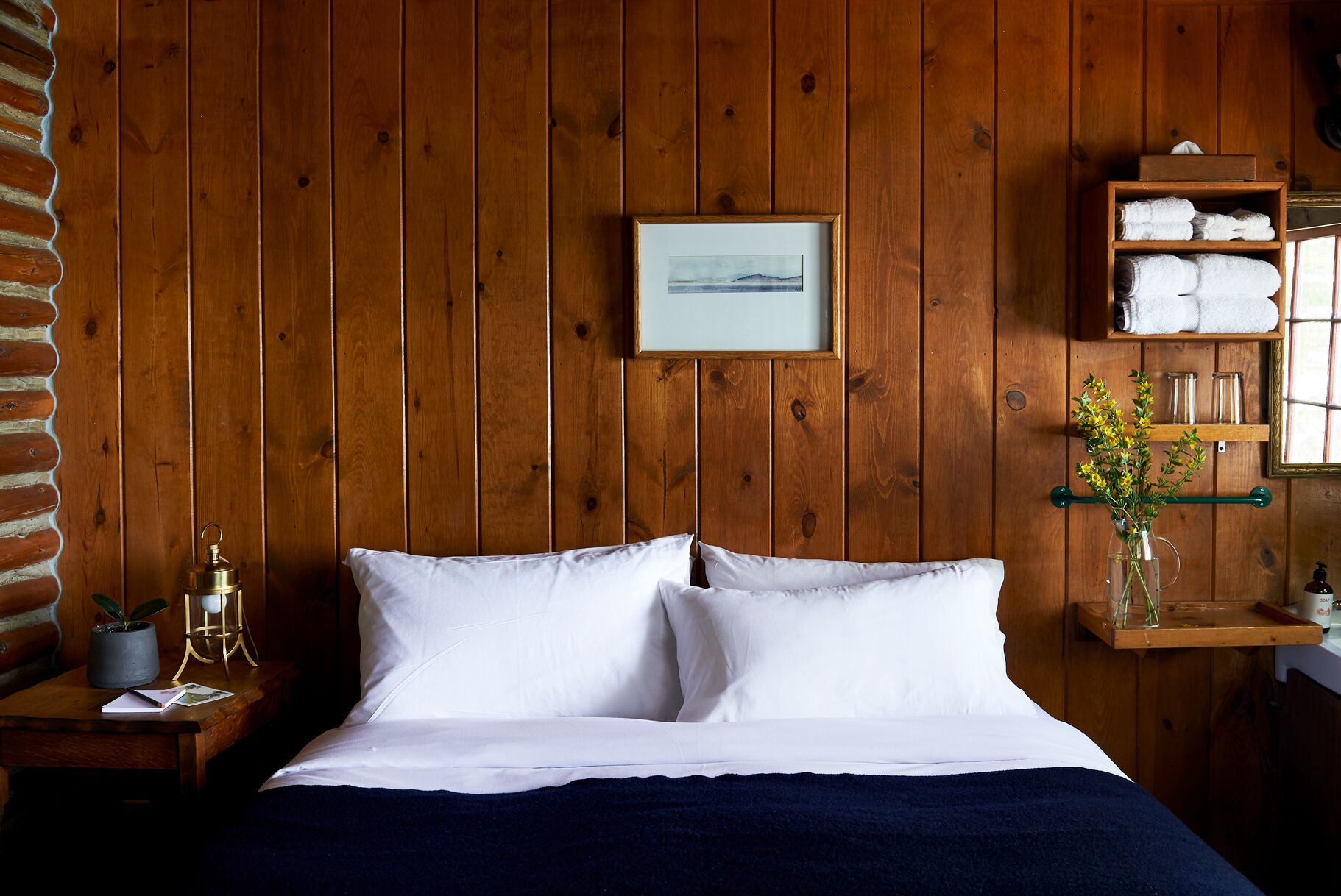 Een kamer met houten lambrisering, een bed, een lijst met een foto, een plank met handdoeken en een vaas met bloemen.