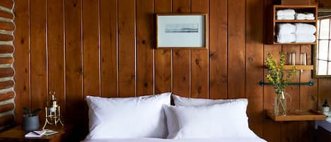 A wooden-paneled room with a bed, a framed picture, a shelf with towels, and a vase with flowers.