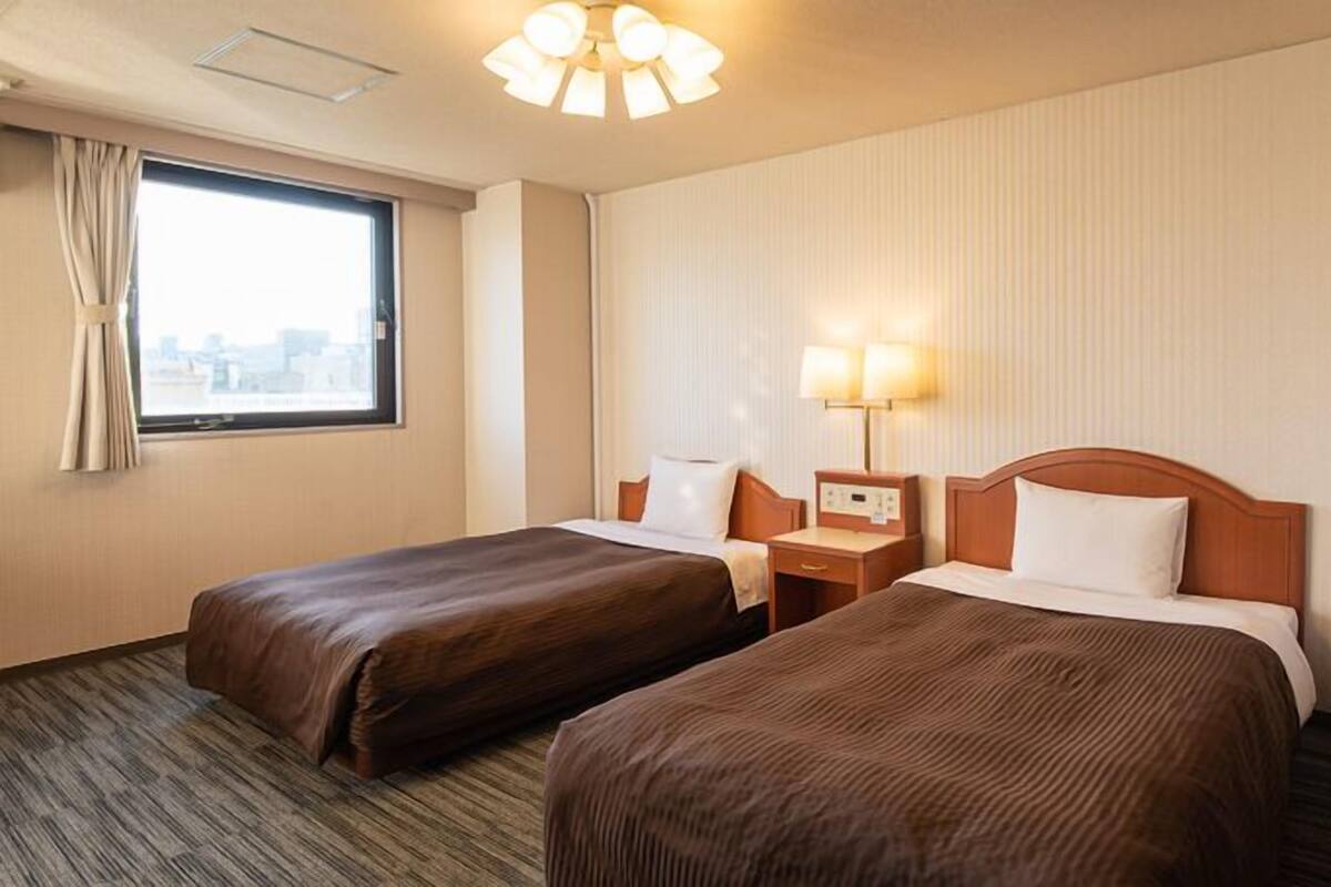 A hotel room with two beds, a window, a lamp, and a ceiling light.