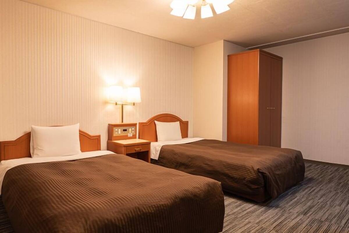 A hotel room with two beds, a nightstand, a lamp, and a wardrobe.