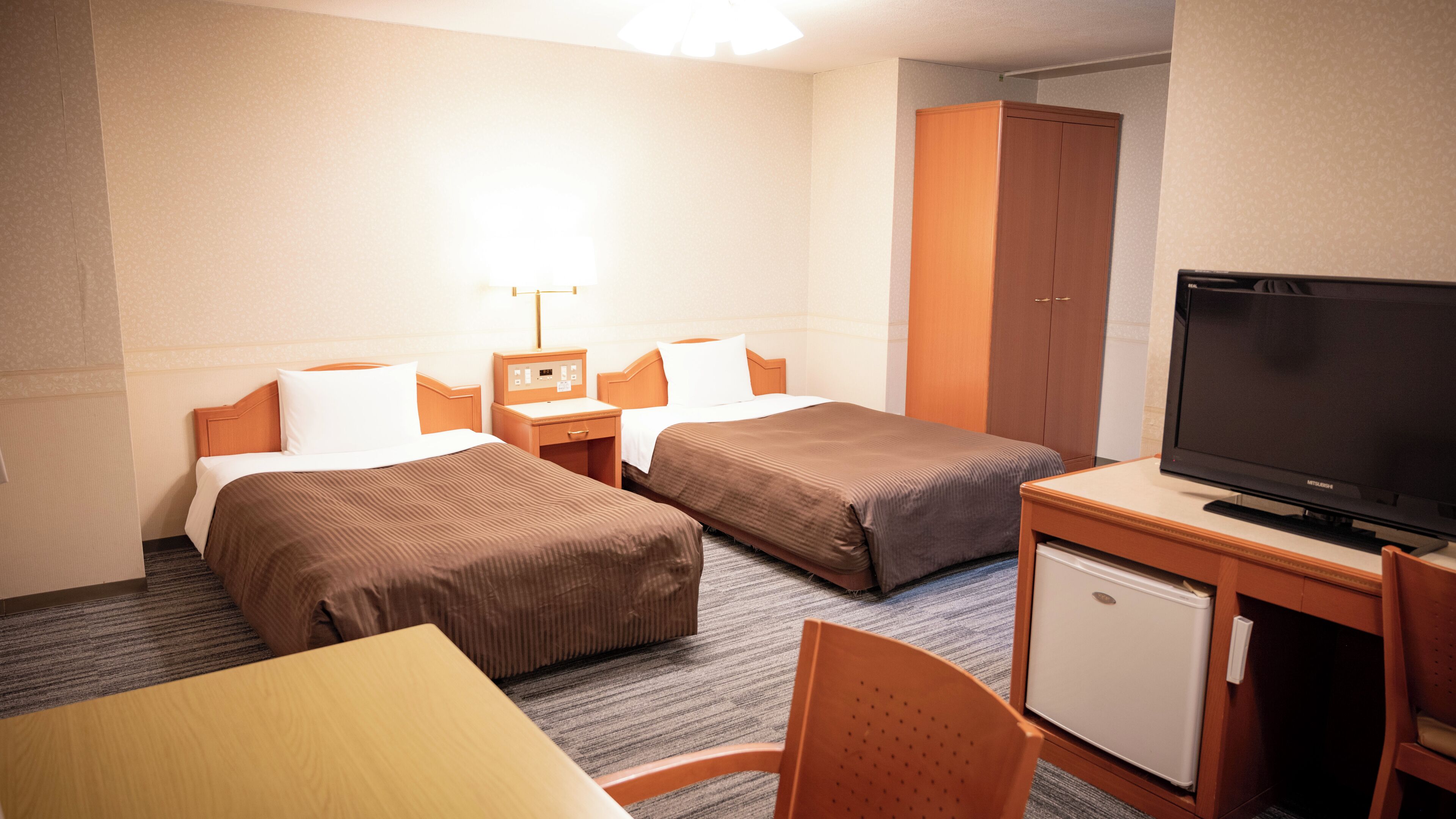 A hotel room with two beds, a desk with a television, a small refrigerator, and a cabinet.