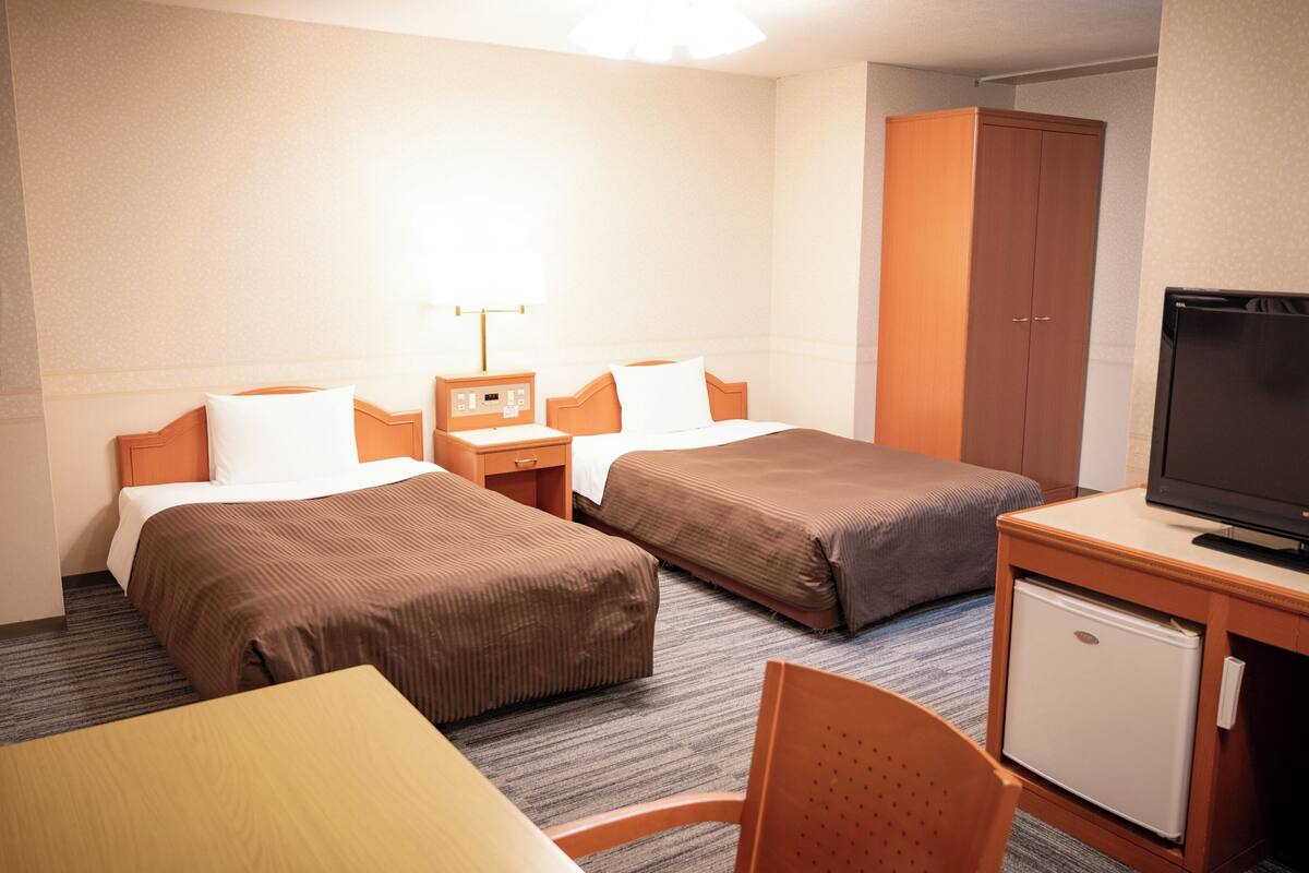A hotel room with two beds, a desk with a television, a small refrigerator, and a cabinet.