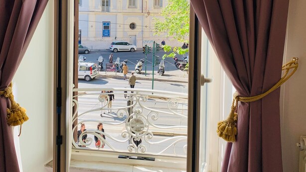A room with a view of a street through an open window, curtains, and a balcony railing. - Trastevere Royal Suite (Rome)